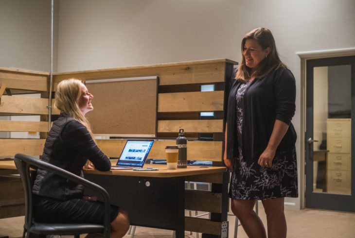 Two people talking while one person sits at their desk working