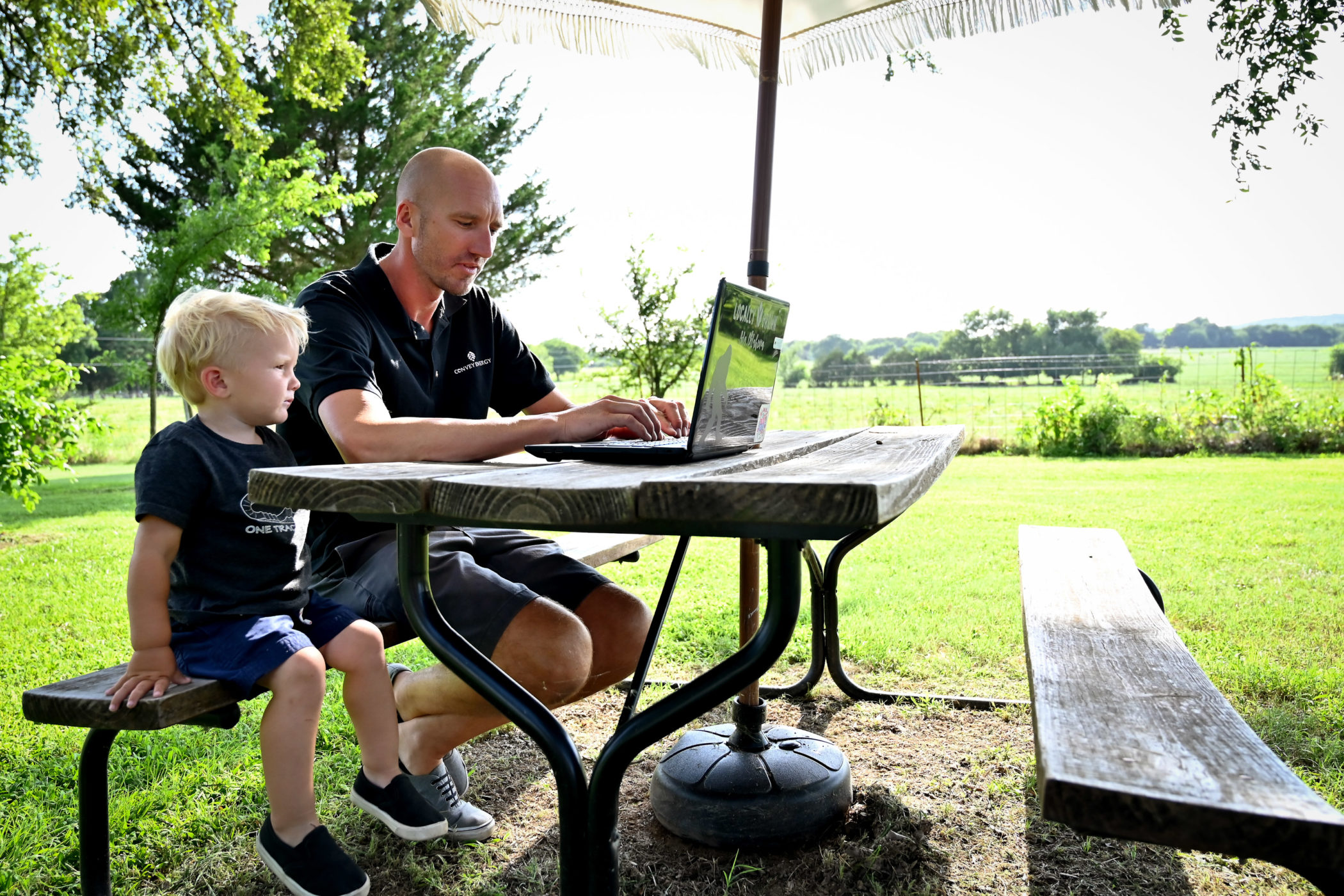 Cody Tucker sits next to his son while he works at a picnic table outside his home