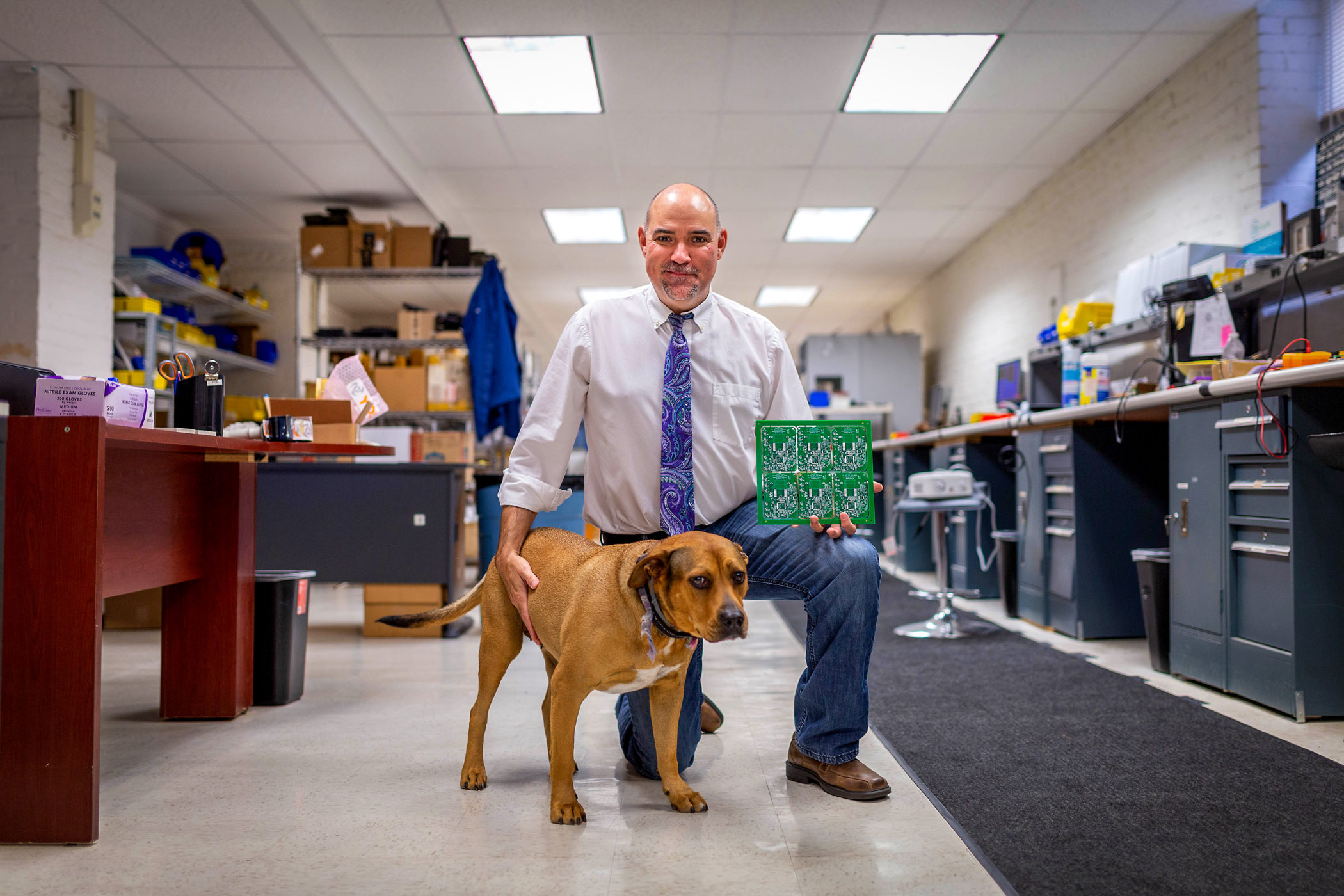 Paul Yost holds a circuit board while petting a dog in his company's workspace
